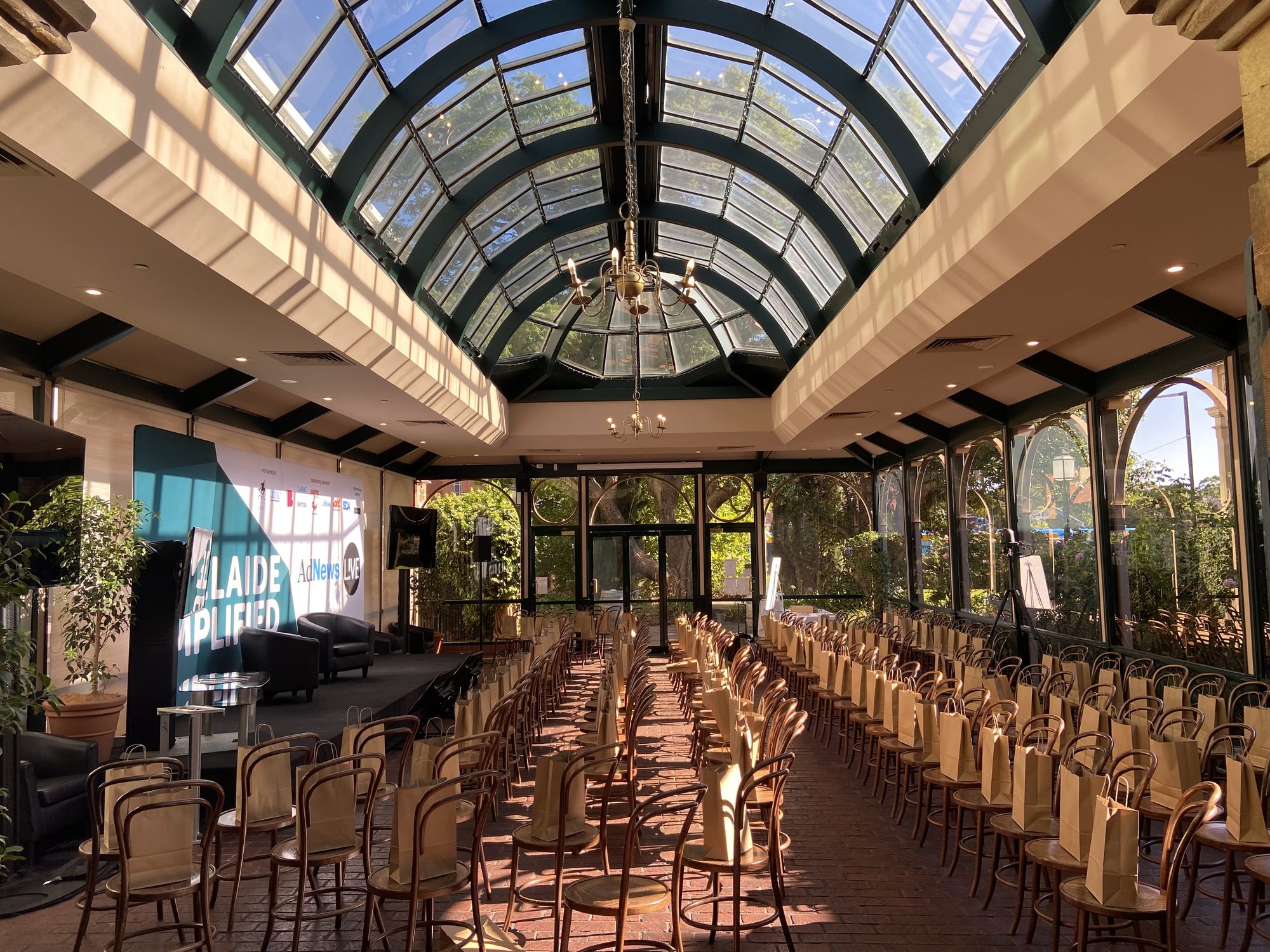 A conference under way in the Conservatory, rows of chairs facing a stage beneath the arched glass roof, morning sun across the room
