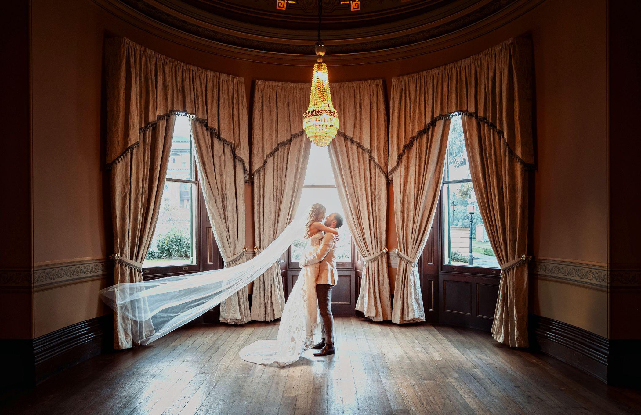 A bride and groom beneath the chandelier in the Ballroom bay window, her veil sweeping the length of the floor