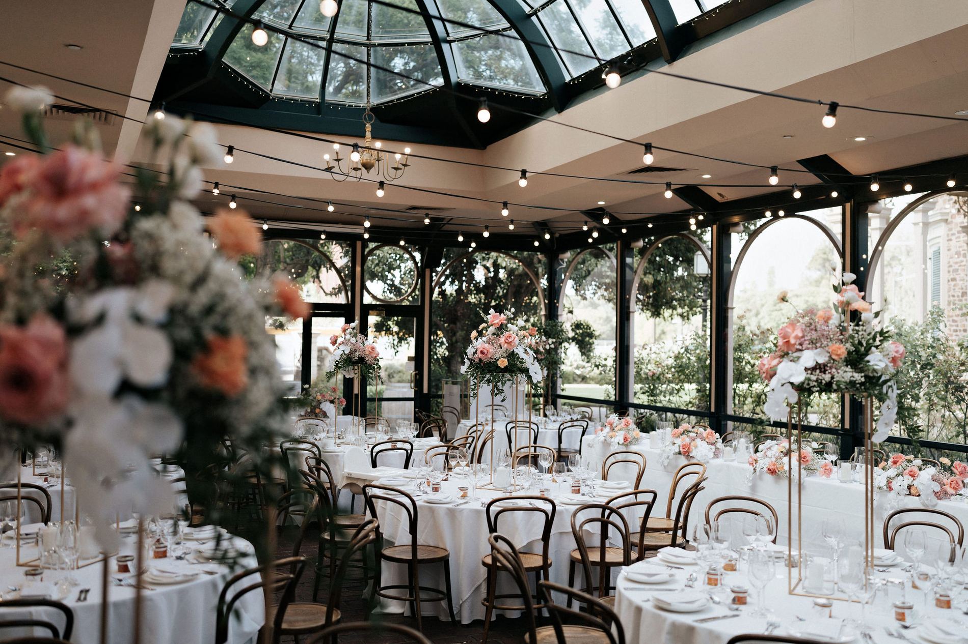 The Conservatory dressed for a wedding, tall coral and white arrangements on gold stands beneath the glass roof