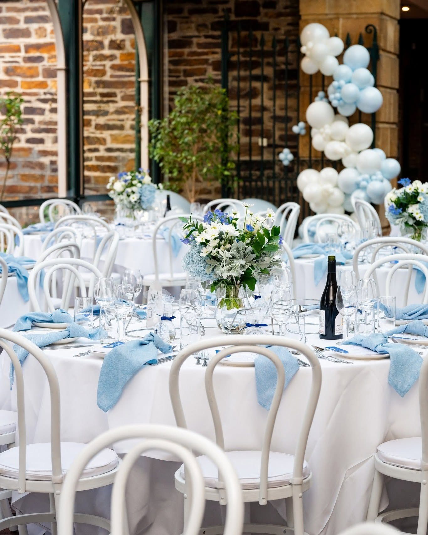 A christening lunch in the Conservatory, round tables with pale blue napkins and a balloon garland by the old stone wall