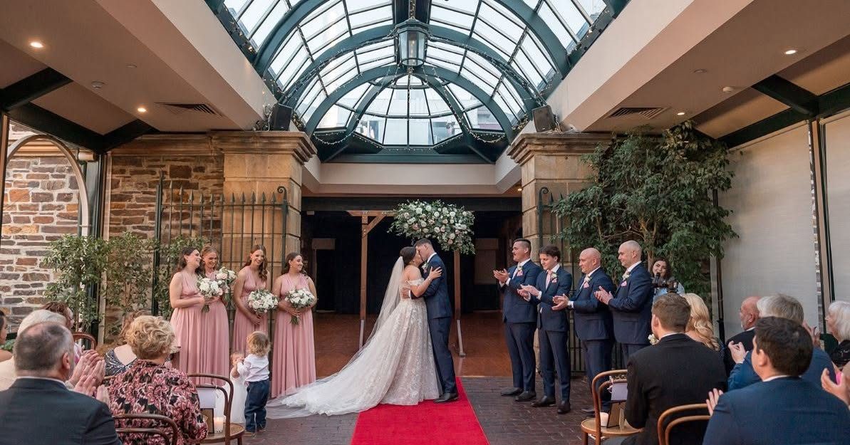 A couple sharing a kiss at their ceremony in the Conservatory, the bridal party either side under the glass dome