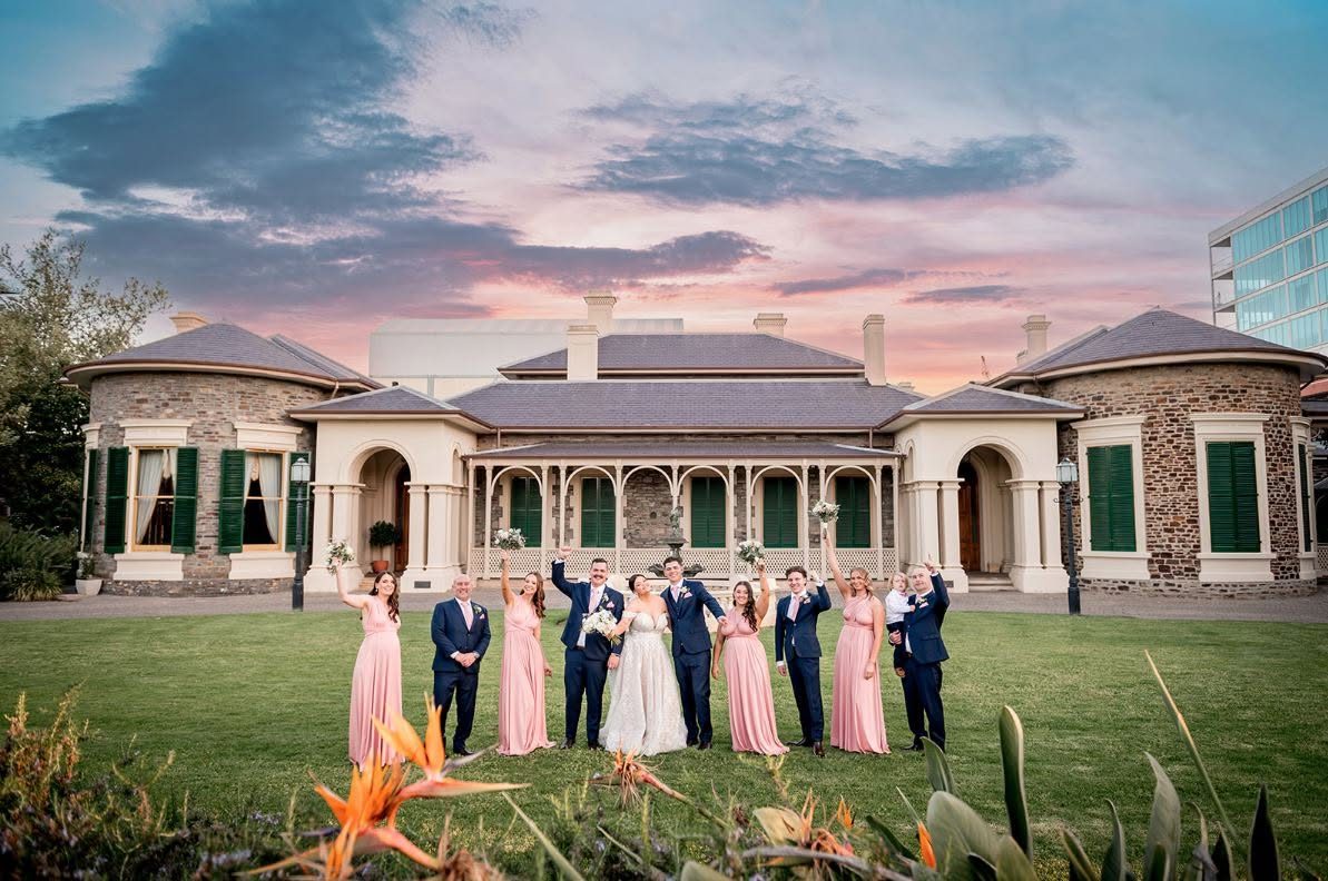 A bridal party celebrating on the front lawn of Ayers House at sunset, the bluestone house behind them