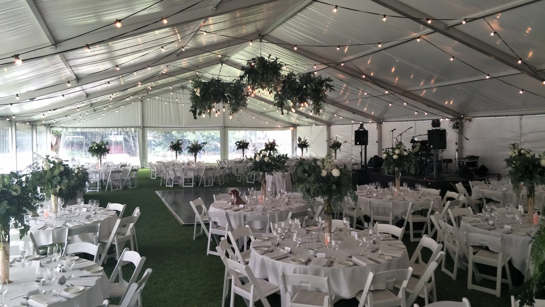 The Marquee set for a wedding, festoon lights and hanging greenery above round tables, a dance floor and a stage for the band