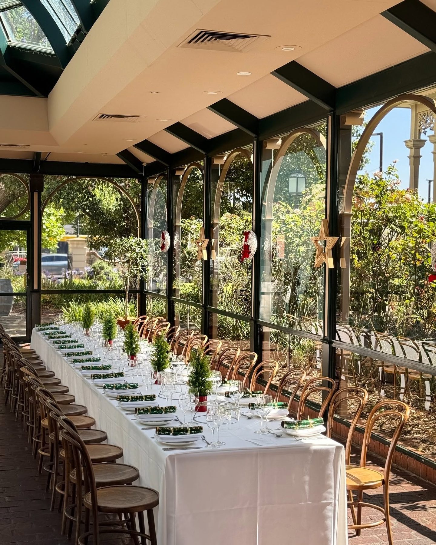 A Christmas long table in the Conservatory, with small trees and crackers down its length and the garden beyond